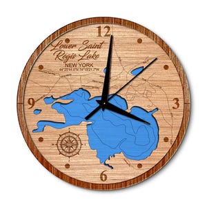 May include: A wooden clock with a map of Lower Saint Regis Lake in New York. The clock face is surrounded by a brown wooden frame. The lake is blue and the surrounding area is brown. The clock hands are black. The clock face also includes the coordinates of the lake: 44°25'44.0"N 74°15'21.7"W.
