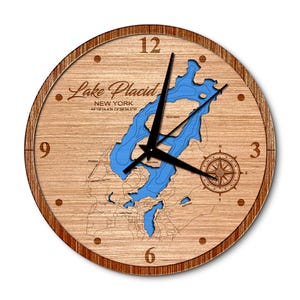 May include: A wooden clock with a blue map of Lake Placid, New York. The clock face has Roman numerals and a compass rose. The text "Lake Placid, New York 44°19'14.4"N 73°58'34.5"W" is printed on the clock face.