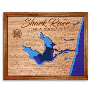 May include: A framed wooden map of Shark River, New Jersey, featuring a light brown wood grain surface with engraved street lines and a compass rose. The water is represented by layered blue cutouts showing varying depths of the river.