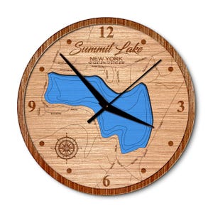 May include: A round wooden wall clock featuring a laser-engraved map of Summit Lake, New York, with coordinates 43°12'37.3"N 73°27'47.0"W. The clock face displays a blue lake cutout, black hands, a compass rose, and brown numerals 3, 6, 9, and 12.