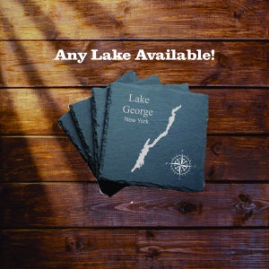 May include: A stack of dark gray slate coasters with a map of Lake George, New York, and a compass rose design. The text "Any Lake Available!" is above the coasters.