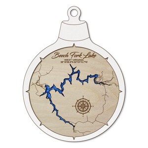 May include: A wooden ornament in the shape of a Christmas bauble, featuring a map of Beech Fork Lake in West Virginia. The lake is depicted in blue against a light brown background, with a compass rose and text that reads "Beech Fork Lake, West Virginia, 38°18'36.8"N 82°23'15.7"W".