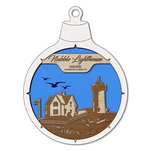 May include: Wooden Christmas ornament featuring a detailed depiction of the Nubble Lighthouse in Maine. The design includes a blue sky, brown buildings, and the text "Nubble Lighthouse, Maine". The ornament is shaped like a Christmas bauble.