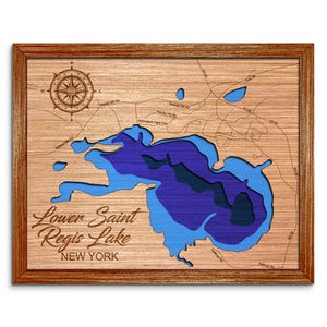 May include: A wooden map of Lower Saint Regis Lake in New York. The lake is shown in shades of blue, with the surrounding land in a light brown color. The map is framed in a dark brown wood frame.
