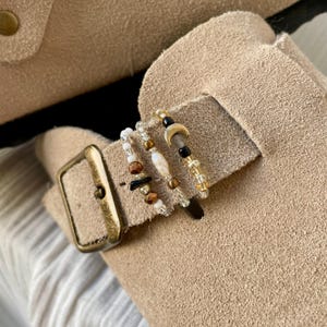 Könnte beinhalten: Drei Perlenringe bestehend aus einer Mischung kleiner weißer, brauner, schwarzer und transparenter Glasperlen. Ein Ring enthält einen kleinen goldfarbenen Halbmond-Anhänger, während ein anderer eine zentrale weiße, ovale Perle aufweist.