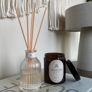 Könnte beinhalten: Eine klare Glasdiffuserflasche mit geriffelten Seiten und Holzdiffuserstäbchen, neben einer braunen Glaskerze mit einem weißen Etikett, auf dem "SIGNATURE  Cire Soy Wax  100 ml | 3.38 oz  Hand Poured in Australia" steht.