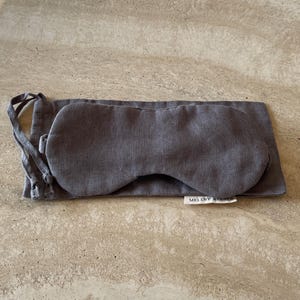 Puede incluir: Una máscara para dormir gris y una bolsa a juego de tela de lino. La máscara para dormir tiene forma ovalada con una correa, y la bolsa es rectangular. La etiqueta de la bolsa dice "MELLBY ATELIER".