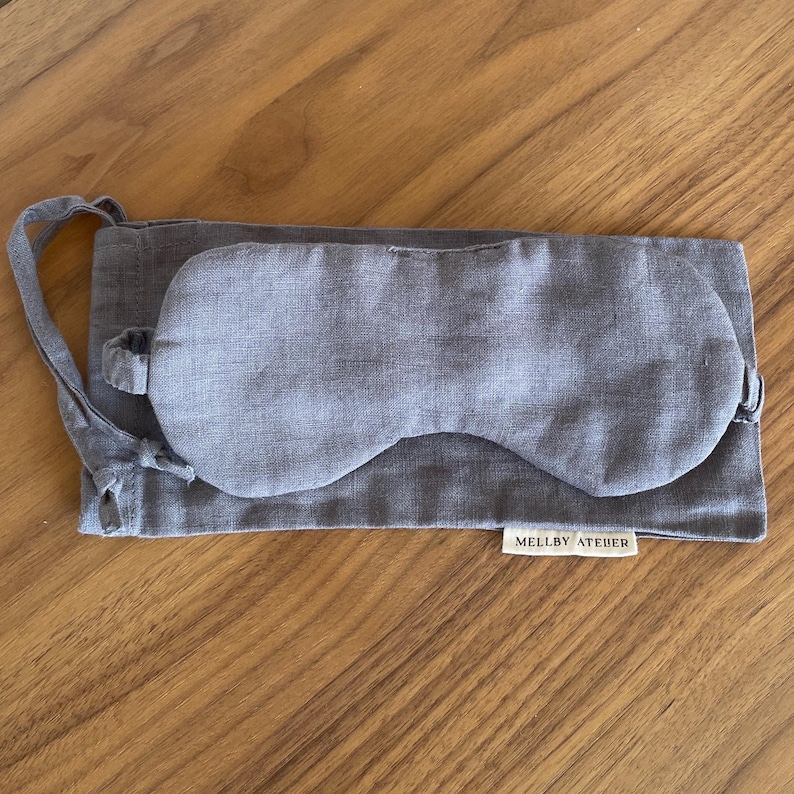 May include: A gray sleep mask and matching pouch made of linen fabric. The mask is oval-shaped with a strap, and the pouch has a drawstring closure. The label on the pouch reads "MELLBY ATEIER".