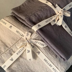 Puede incluir: Dos juegos de ropa de cama de lino, uno gris y otro beige, están atados con una cinta blanca. La cinta tiene el texto "MELLBY ATELIER" impreso en ella.