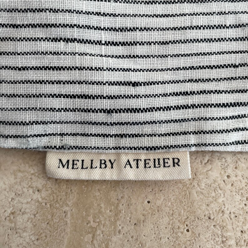 Closeup of the Mellby Atelier tag on a soft ivory/black pinstripes sleep mask drawstring bag.