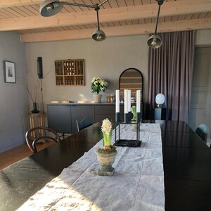 May include: A dining room featuring a long black table with a light grey linen runner, a white hyacinth in a weathered stone urn, and a black metal candelabra with four white candles. A dark sideboard, a wooden wall display case, and a ceiling light fixture.