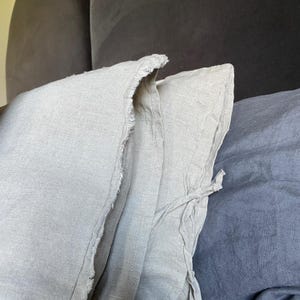 May include: Two light beige linen pillowcases featuring a frayed edge detail and a side tie closure. The fabric has a visible woven texture, and the pillowcases are positioned next to a dark grey linen pillowcase.