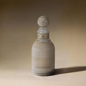 Könnte beinhalten: Eine klare Glasflasche mit einer strukturierten Oberfläche und einem runden Glasstopfen. Die Flasche hat ein Etikett mit dem Text "MER" und einem Dreieckslogo.