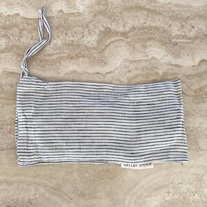 Mellby Atelier linen sleep mask in soft ivory/black pinstripes, shown in its drawstring bag and displayed on a stone counter.