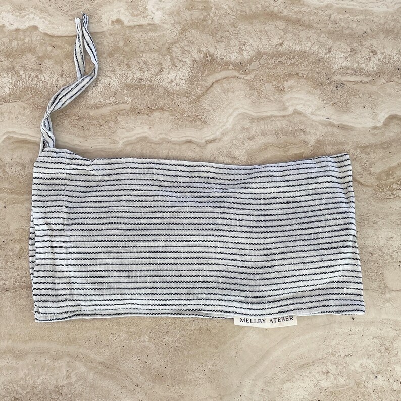 Mellby Atelier linen sleep mask in soft ivory/black pinstripes, shown in its drawstring bag and displayed on a stone counter.