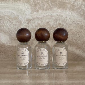 Puede incluir: Tres botellas de spray ambiental de vidrio transparente con tapas esféricas de madera marrón oscuro. Cada botella tiene una etiqueta ovalada color crema con texto negro que dice Signature, Apple Orchard y Pink Grapefruit, más detalles.