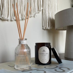 Vanilla-scented coconut soy candle in amber jar with black lid, styled beside a clear glass reed diffuser on a modern neutral-toned sideboard.