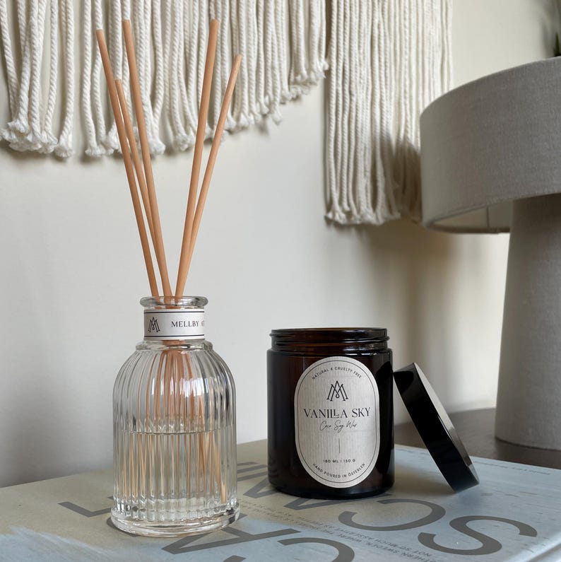 Vanilla-scented coconut soy candle in amber jar with black lid, styled beside a clear glass reed diffuser on a modern neutral-toned sideboard.