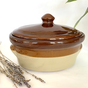 May include: A brown and cream ceramic casserole dish with a lid. The dish has two handles on the sides.