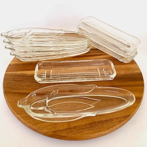 May include: A set of vintage glass serving dishes, including a stack of rectangular dishes and two corn-shaped dishes. The dishes are clear glass with a textured surface and are displayed on a wooden tray.