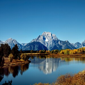 May include: A scenic view of a mountain range with snow-capped peaks reflected in a still lake. The foreground features a grassy bank with trees and bushes in shades of green, yellow, and brown.