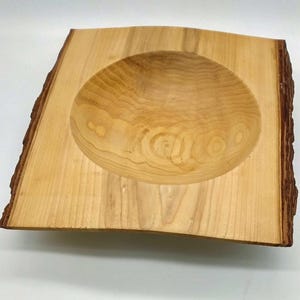 May include: A wooden bowl with a natural edge. The bowl is carved from light-coloured wood, featuring a rounded interior and a rectangular base. The wood grain is visible, adding to the rustic charm of this decorative piece.