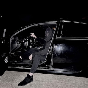 May include: A person wearing a black hooded sweatshirt and black trousers sits in the driver's seat of a black car with the door open. The person's face is covered by a black mask.