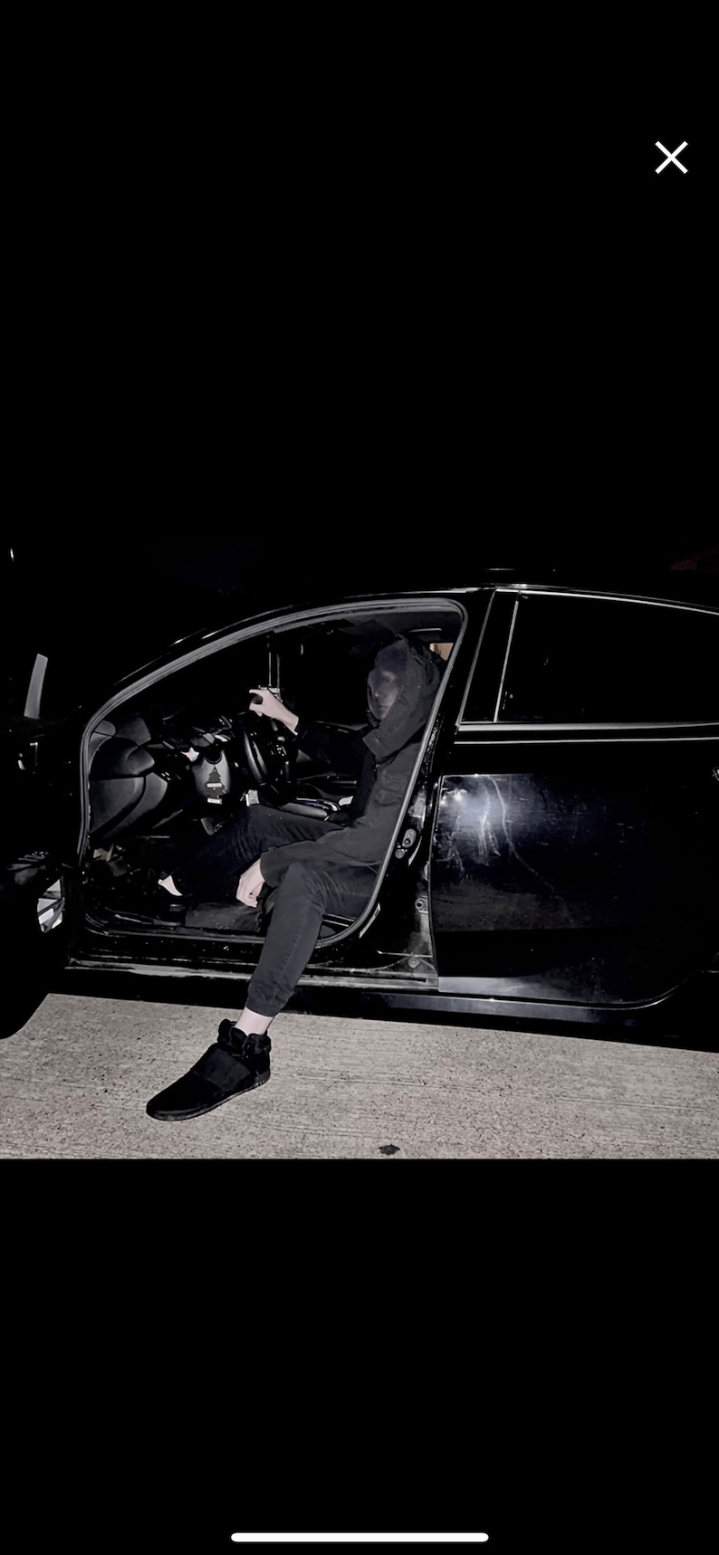 May include: A person wearing a black hooded sweatshirt and black trousers sits in the driver's seat of a black car with the door open. The person's face is covered by a black mask.
