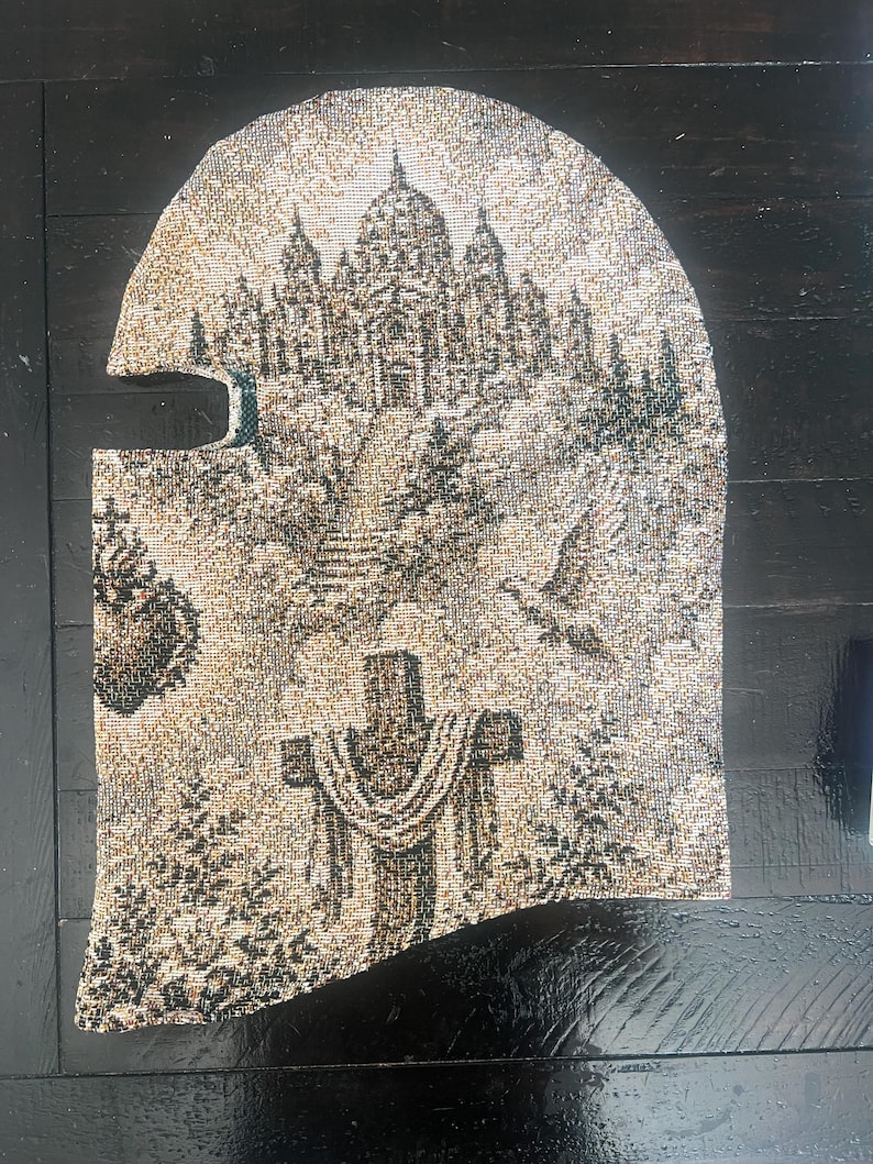 May include: A woven balaclava featuring a detailed tapestry-style design in shades of beige, brown, and black. The pattern depicts a large domed building on a hill, a cross draped with fabric, a flying bird, and a heart-shaped emblem on the side.