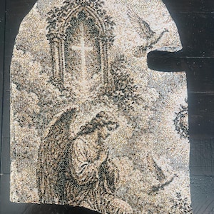 May include: A woven balaclava featuring a detailed tapestry-style pattern in shades of black, beige, and white. The design depicts a kneeling angel with wings positioned below a gothic archway containing a cross, with clouds in the background.