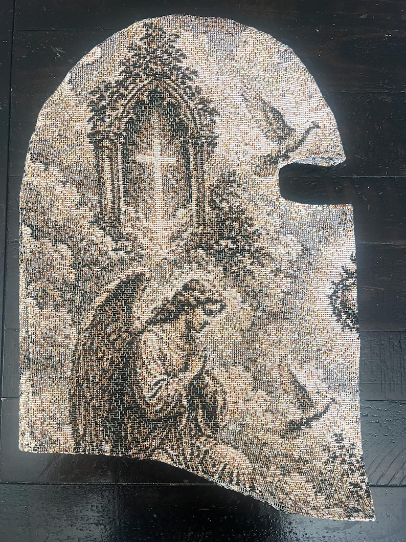 May include: A woven balaclava featuring a detailed tapestry-style pattern in shades of black, beige, and white. The design depicts a kneeling angel with wings positioned below a gothic archway containing a cross, with clouds in the background.