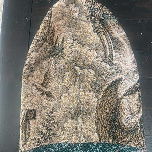 May include: A slouchy beanie hat crafted from a woven tapestry fabric featuring a detailed landscape scene with trees, birds, and architectural elements in shades of gold, brown, and black, with a contrasting dark teal green interior lining.