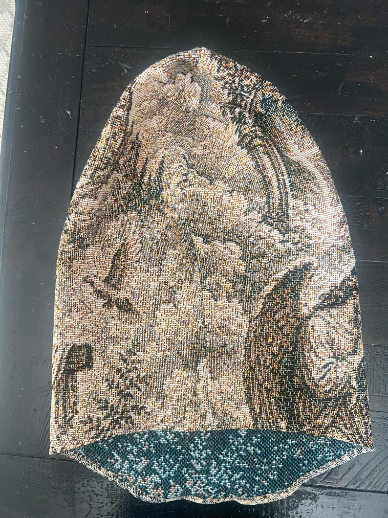 May include: A slouchy beanie hat crafted from a woven tapestry fabric featuring a detailed landscape scene with trees, birds, and architectural elements in shades of gold, brown, and black, with a contrasting dark teal green interior lining.