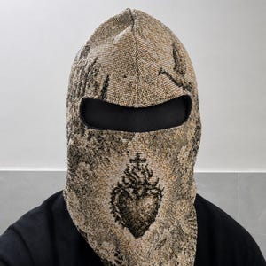 May include: A woven balaclava featuring a beige and black tapestry-style pattern with a prominent sacred heart emblem centered on the front. The headgear includes a single horizontal eye opening lined with black fabric, covering the head and neck.