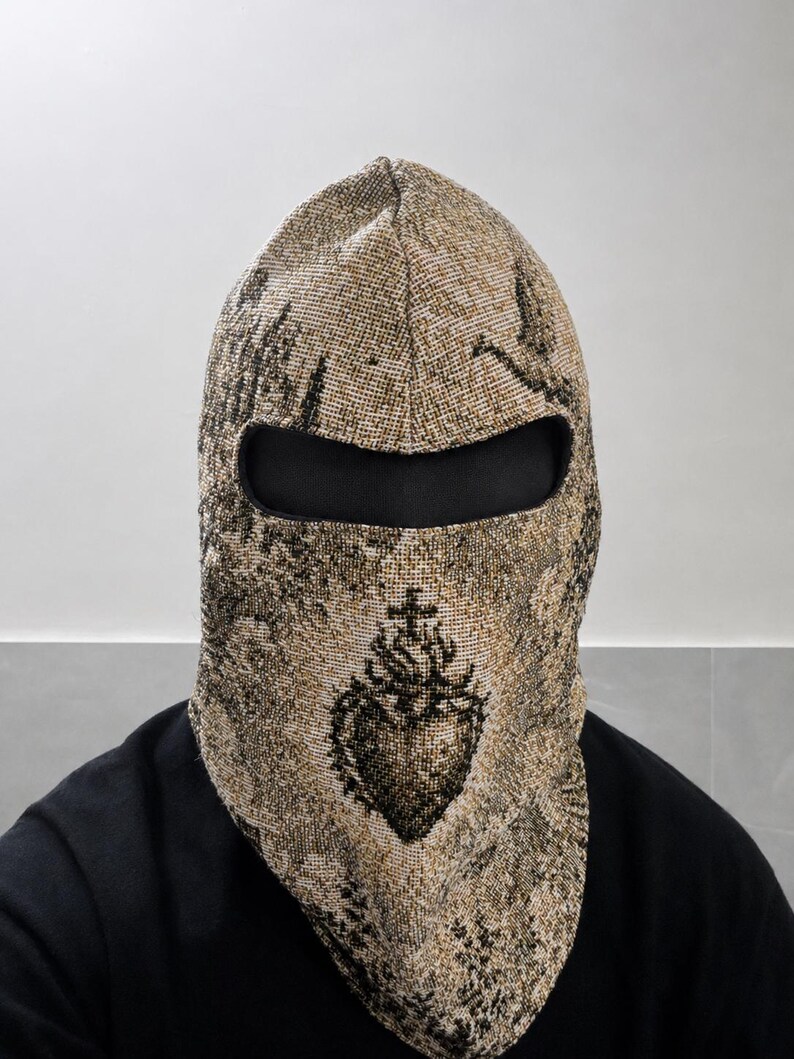 May include: A woven balaclava featuring a beige and black tapestry-style pattern with a prominent sacred heart emblem centered on the front. The headgear includes a single horizontal eye opening lined with black fabric, covering the head and neck.