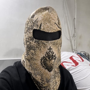 May include: A woven balaclava featuring a textured gold and black pattern with a prominent black sacred heart emblem centered on the front. The headgear includes a single horizontal eye opening and covers the head and neck with a thick fabric.