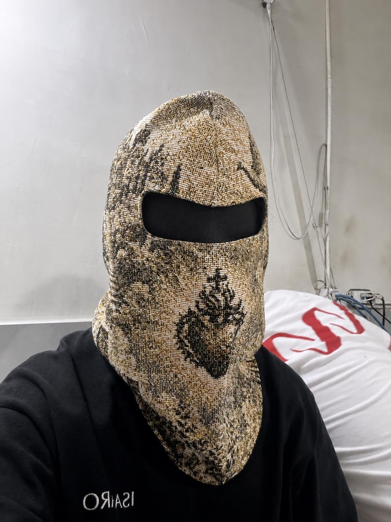 May include: A woven balaclava featuring a textured gold and black pattern with a prominent black sacred heart emblem centered on the front. The headgear includes a single horizontal eye opening and covers the head and neck with a thick fabric.