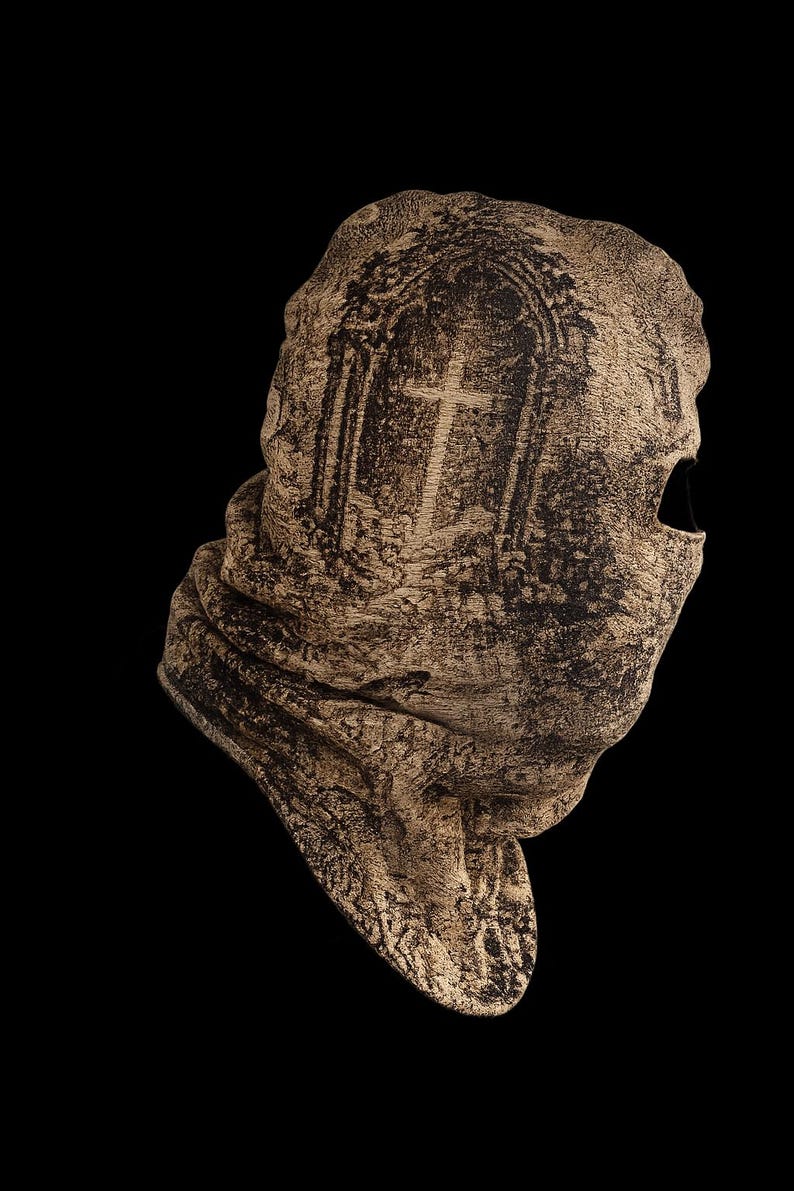 May include: A beige and black textured balaclava featuring a printed graphic of an arched doorway with a cross inside. The fabric has a weathered, stone-like appearance with a single eye opening on the right side and a folded neck section.