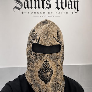 May include: A tan and black woven balaclava featuring a detailed graphic of a sacred heart with a cross on top. The fabric has a textured appearance with a single eye opening. The text Saints Way Forged By Faith Est. 2026 is visible in the background.
