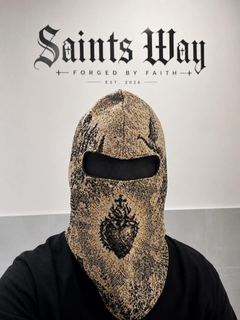 May include: A tan and black woven balaclava featuring a detailed graphic of a sacred heart with a cross on top. The fabric has a textured appearance with a single eye opening. The text Saints Way Forged By Faith Est. 2026 is visible in the background.