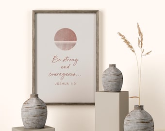 May include: A framed print with the text "Be strong and courageous..." and a pink sun design. Two decorative vases with a textured, gray finish sit on pedestals. A sprig of dried grass adds a natural element to the display. The overall theme is inspirational home decor.