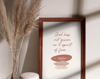 May include: A framed print with the text "God has not given us a spirit of fear..." and the scripture reference 2 Timothy 1:7. The frame is brown, and the print has a neutral color palette. A vase with dried plants and a white jar are in the foreground.
