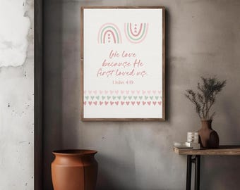 May include: A rectangular wall art print in a wooden frame featuring two pastel rainbows at the top, the text "We love because He first loved us. 1 John 4:19" in pink script, and a repeating pattern of small pink and green hearts at the bottom.