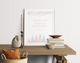 May include: A framed wall art print featuring the text God has not given us a spirit of fear 2 Timothy 1:7 in pink script, with a row of pastel pink, blue, and yellow triangles at the bottom, displayed in a white frame on a wooden surface.