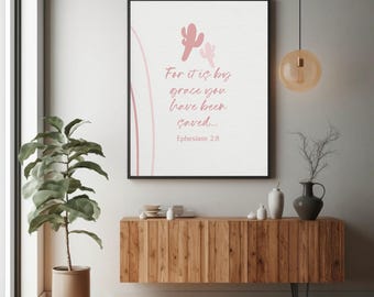 May include: A rectangular wall art print in a black frame featuring a white background with pink cactus illustrations and the text: For it is by grace you have been saved... Ephesians 2:8. The design includes soft pink curved line accents.
