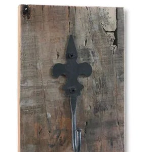 Könnte beinhalten: Ein rustikaler Wandhaken mit einem Fleur-de-Lis-Design. Der schwarze Metallhaken ist auf einem rechteckigen Stück Altholz montiert. Das Holz hat ein verwittertes Aussehen mit verschiedenen Braun- und Grautönen.