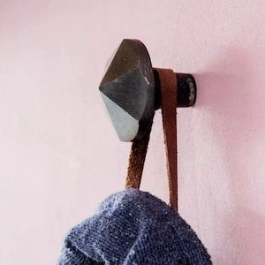 May include: A black diamond-shaped hook with a brown leather strap hanging from it. The hook is mounted on a pink wall.