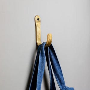 May include: A gold metal hook mounted on a grey wall with two blue denim tote bags hanging from it.