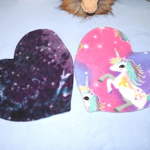May include: Two heart-shaped fabric pieces. One piece features a dark purple and black galaxy print with small white stars. The other piece displays a pink and purple background with a white unicorn, colorful mane, and star patterns.
