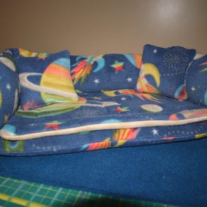 May include: A small, plush sofa bed featuring a dark blue fleece fabric with a colorful space-themed print of planets, stars, and rockets. The sofa includes two matching square throw pillows and a removable seat cushion with a light-colored trim.